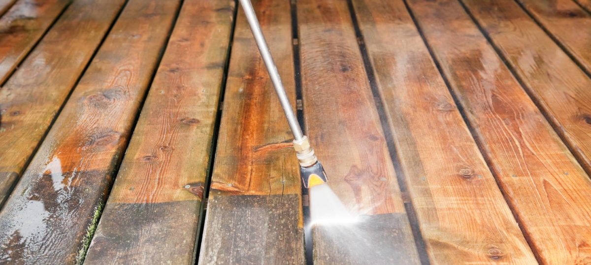 A closeup of a professional power washer's pressure washing wand spraying a weathered wooden deck to expose the fresh redwood underneath