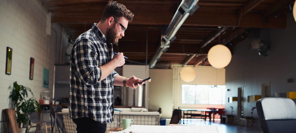 A bearded architect in a blue and white plaid shirt standing at a drafting table checking his Insurance policy on his phone