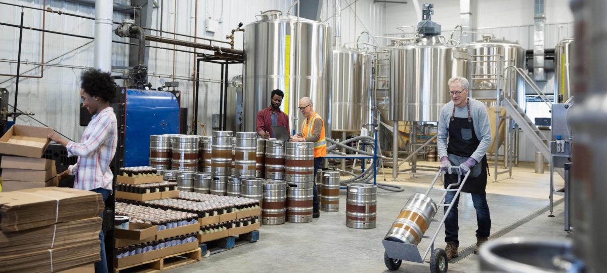 Man wheeling a beer keg on a stainless steel dolly through a brewery with three other employees, a row of kegs, and stainless steel fermentation tanks in the background