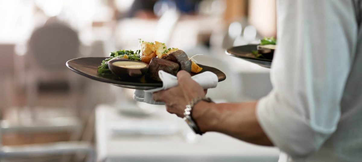 Closeup of catering business owner in a white dress shirt with rolled up sleeves carrying two elegantly plated diners with filet medallions, a small salad, soup and fried potatoes