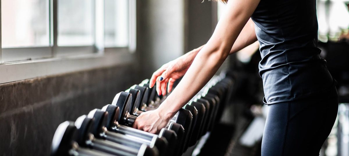 Closeup of a woman in a dark tank top picking up dumbbells from a rack in a fitness center