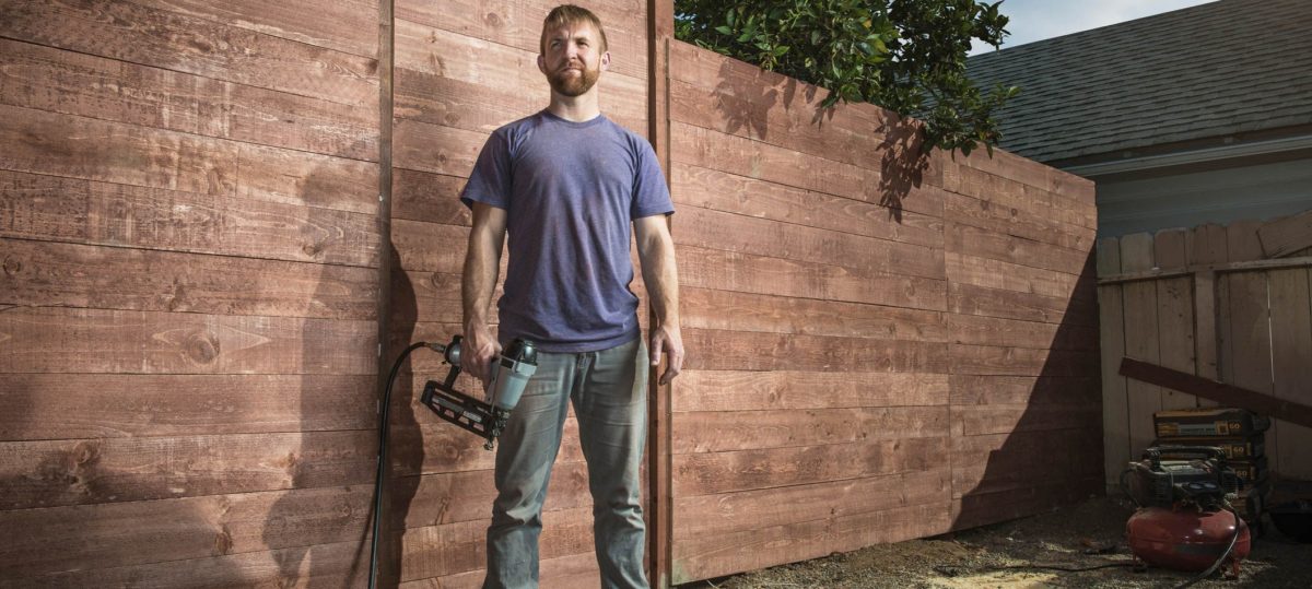 Handyman in dirty purple shirt and blue jeans holding a nail gun in front of a recently finished wooden fence he built