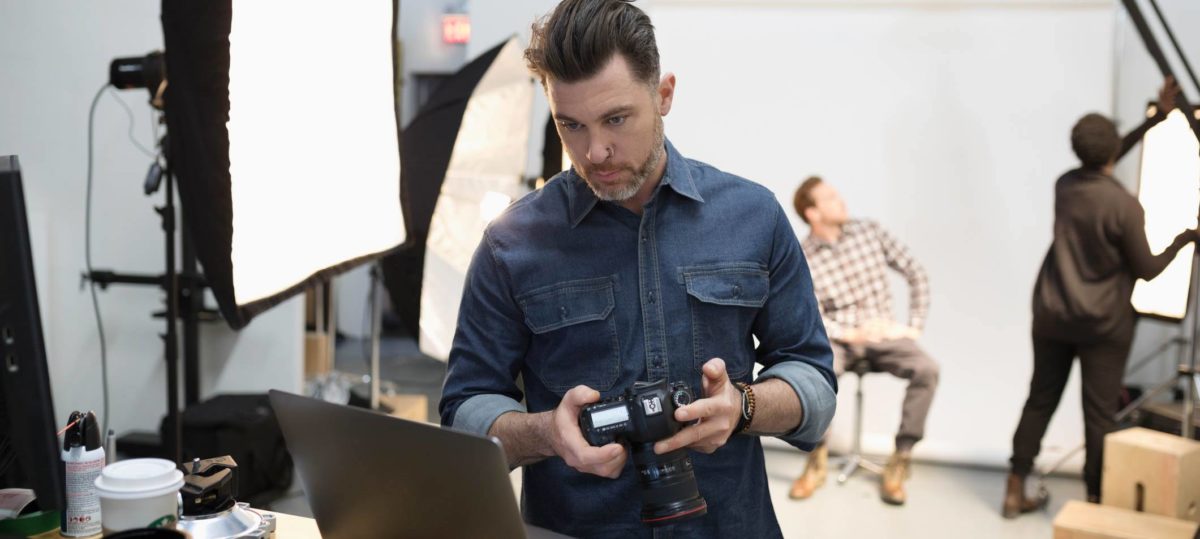 Male photographer holding camera digital camera in front of a laptop at photo shoot with a man in a plaid shirt in the background