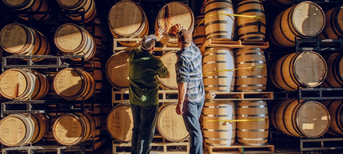 Two men in baseball hats examining barrel of wine at a vineyard