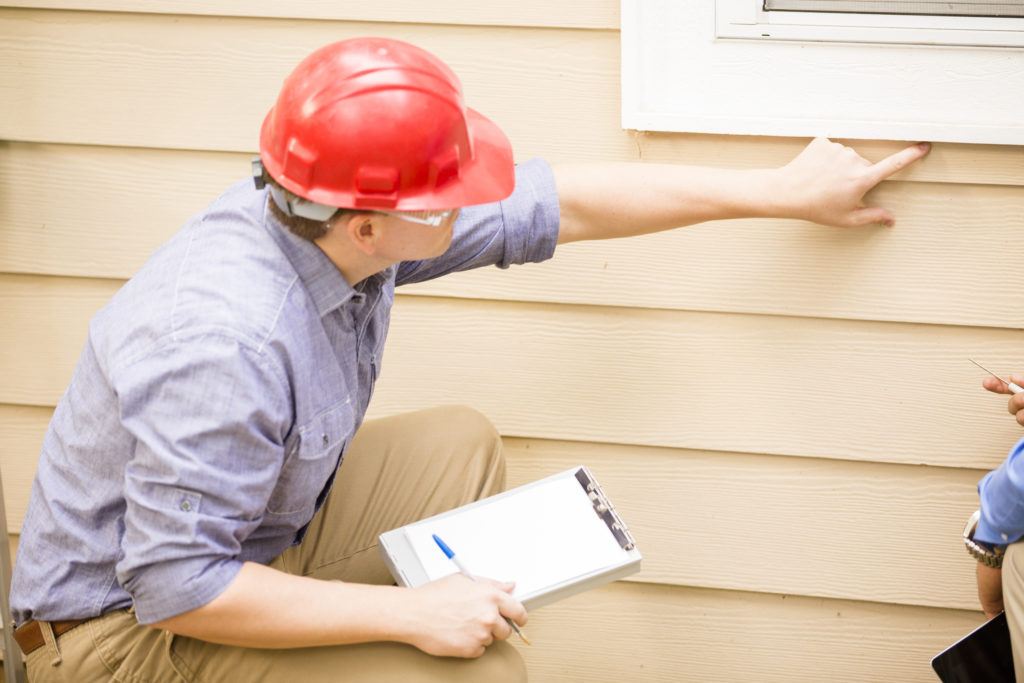Home inspector in blue collared shirt and red hard hat holding a clipboard while inspecting the siding of a house